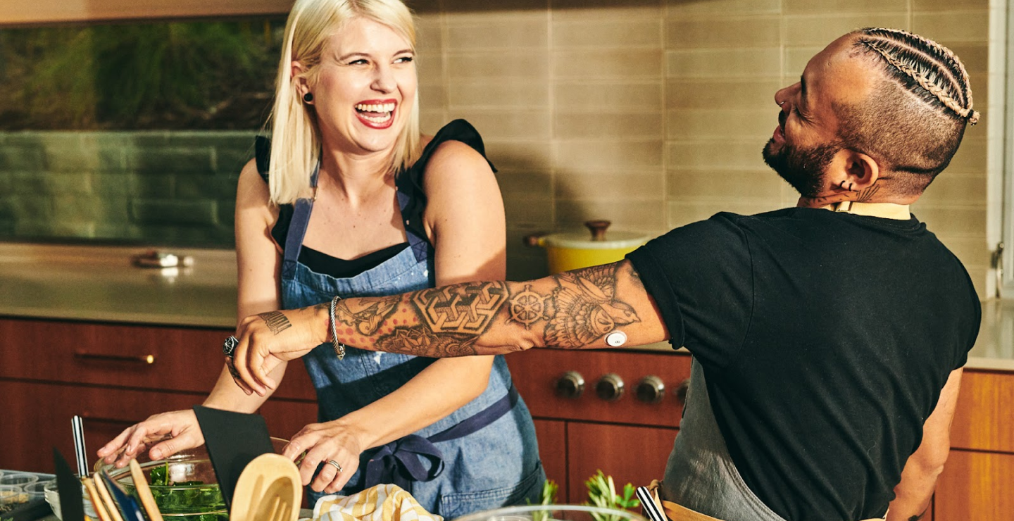 A cheerful couple, one with a glucose monitor patch, happily cooking together in a kitchen.